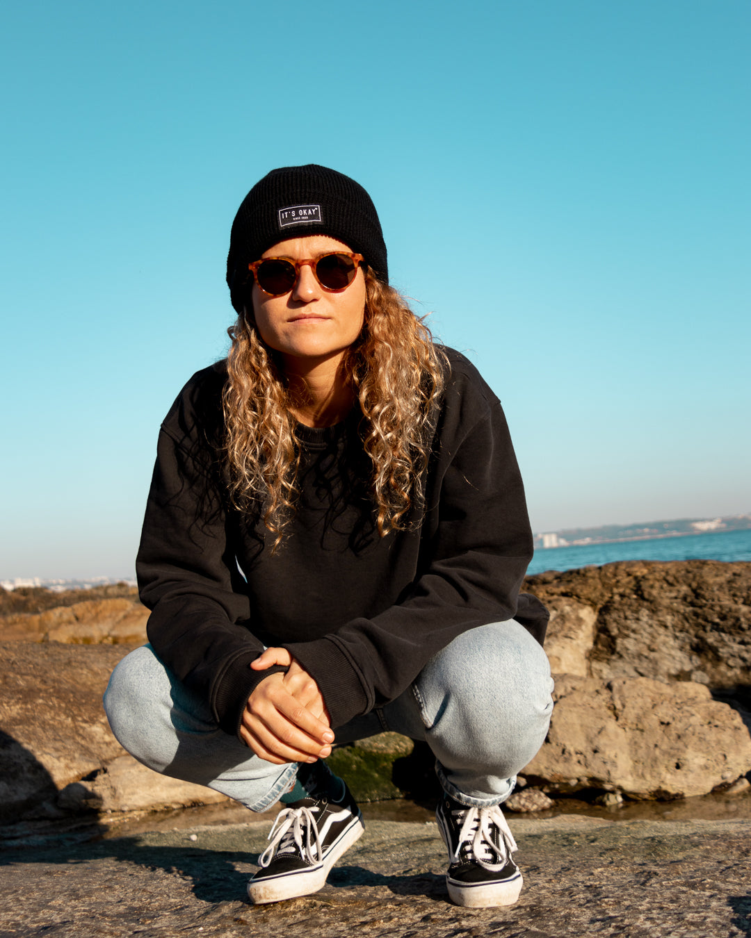 Black Beanie from It's okay | made in Portugal with love | feel cozy and warm with this knitted beanie. Pt gorro de inverno it's okay em preto. Image: women in squat position near the beach wearing black waffle beanie.